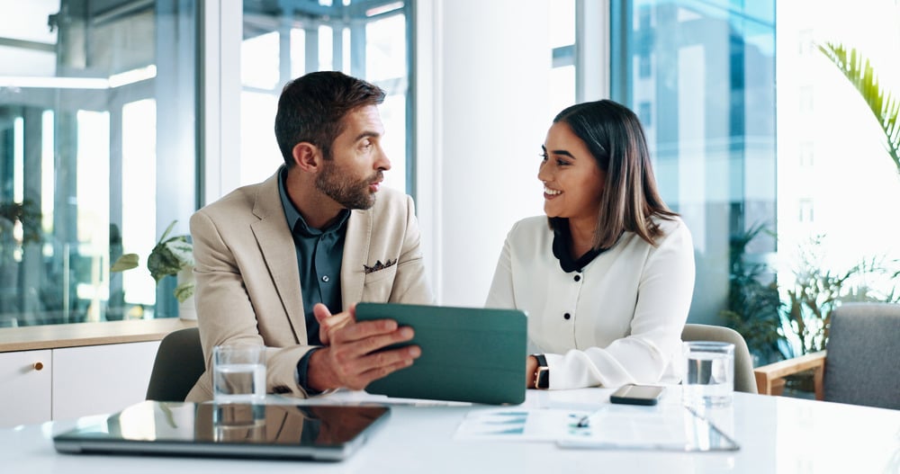 Smiling woman sits with businessman in brightly lit room reviewing tablet