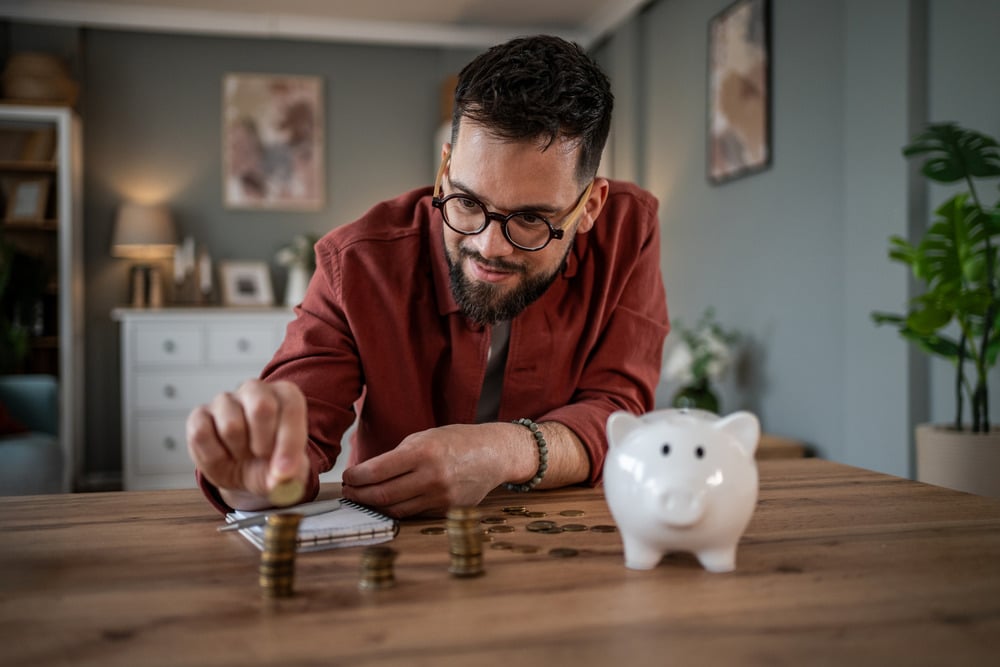 Man budgeting finances and counting stacked coins on a wooden table next to a white piggy bank, indicating personal investment, smart spending, and financial planning at home